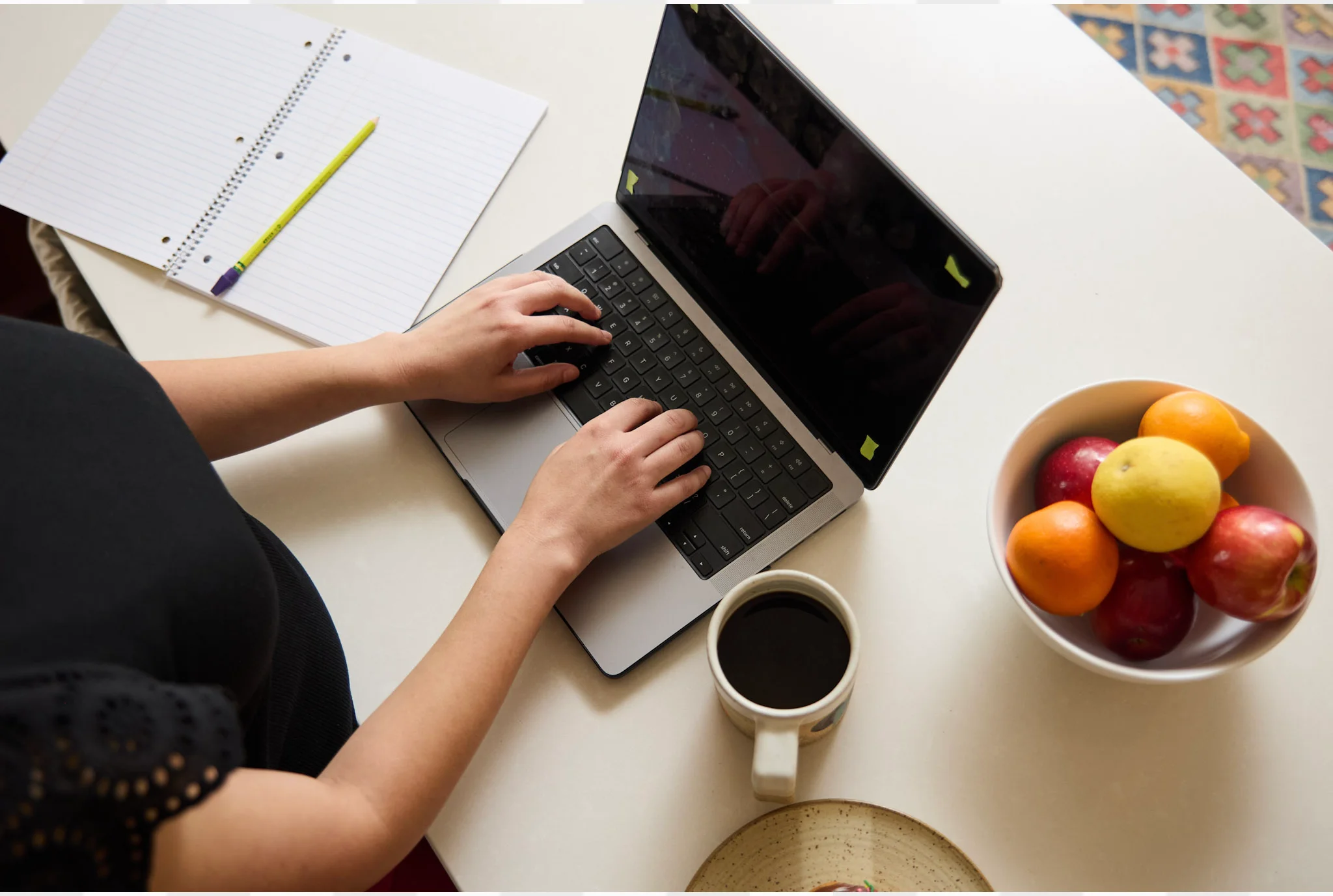 Una persona escribe en una computadora portátil en un escritorio blanco con un cuaderno, lápiz, taza de café y un tazón de fruta, creando un ambiente productivo y acogedor.