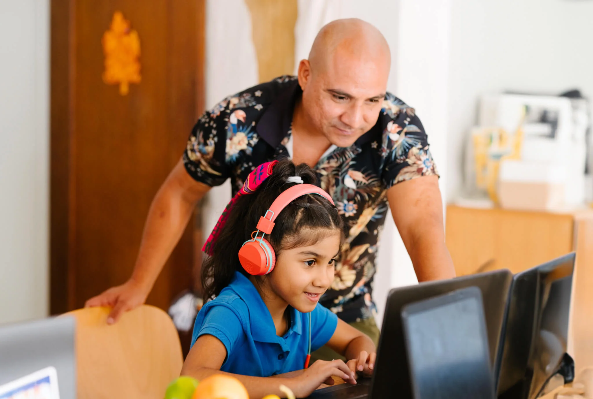 Una niña sonriente con auriculares rojos se sienta en un escritorio usando una computadora, mientras que un hombre con una camisa floral se inclina, mirando con apoyo.