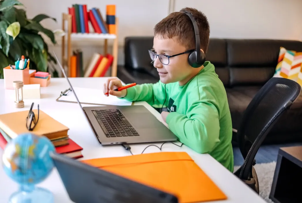 Un niño con gafas y auriculares, vestido con una camisa verde, estudia atentamente en una computadora portátil en un escritorio blanco. Libros, un globo terráqueo y un sofá están en el fondo.