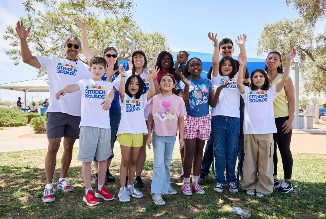 Un grupo diverso de personas sonrientes, adultos y niños, posan al aire libre con los brazos levantados bajo los árboles. Todos llevan camisetas de "Strider Squads", exudando alegría.