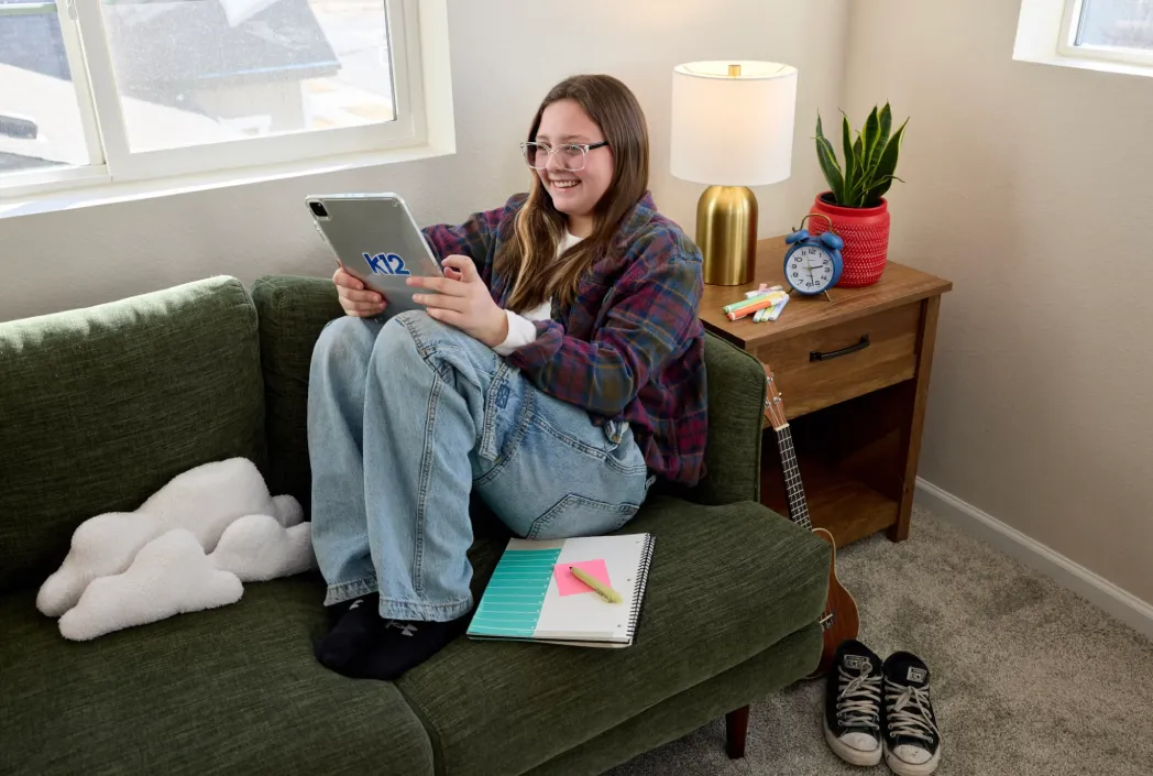 Una persona sonriente con gafas se sienta en un sofá verde, sosteniendo una tableta etiquetada como "K12". Cerca hay un cuaderno, una lámpara, un reloj y una guitarra, que transmiten un ambiente acogedor y estudioso.