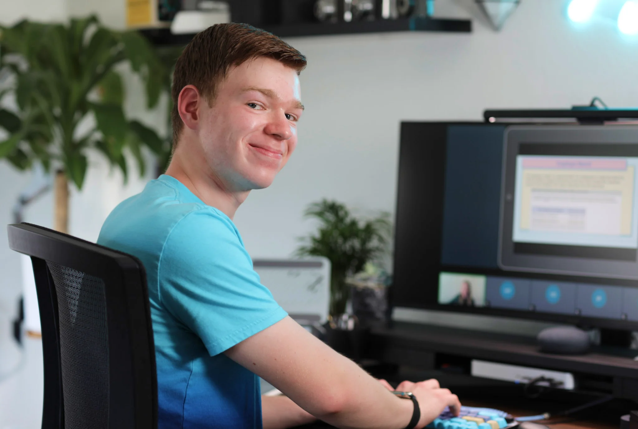 Adolescente con una camisa azul sonríe mientras está sentado en un escritorio con una computadora que muestra una videollamada y una presentación. Planta verde en el fondo.