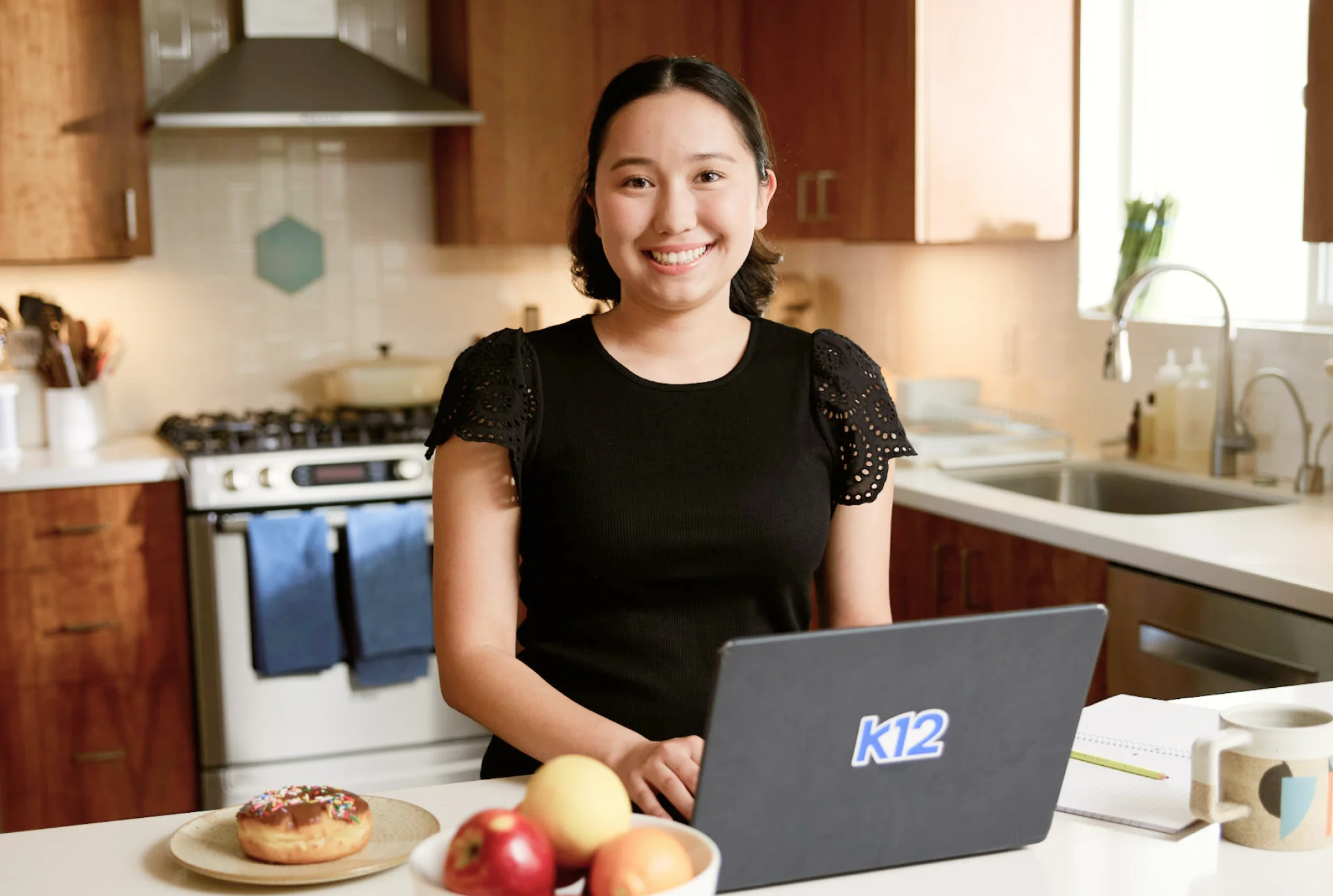 Una mujer está de pie sonriendo en una cocina usando una computadora portátil con "K12" en ella. Un plato con una rosquilla y manzanas está en primer plano, creando una escena cálida y acogedora.