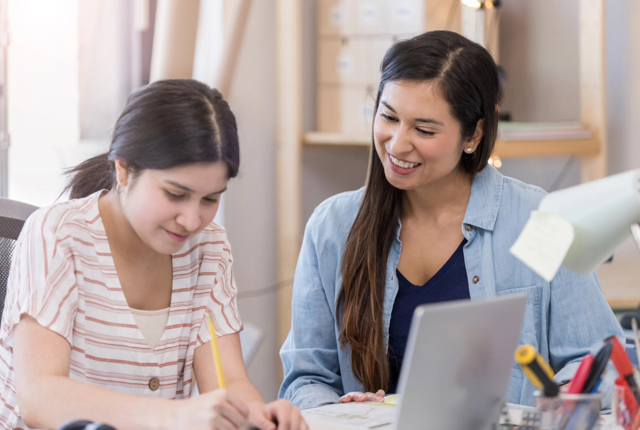 Una mujer sonríe de apoyo mientras una niña más joven escribe en un escritorio, sugiriendo una escena de tutoría o tutoría. Una computadora portátil y materiales de estudio son visibles.