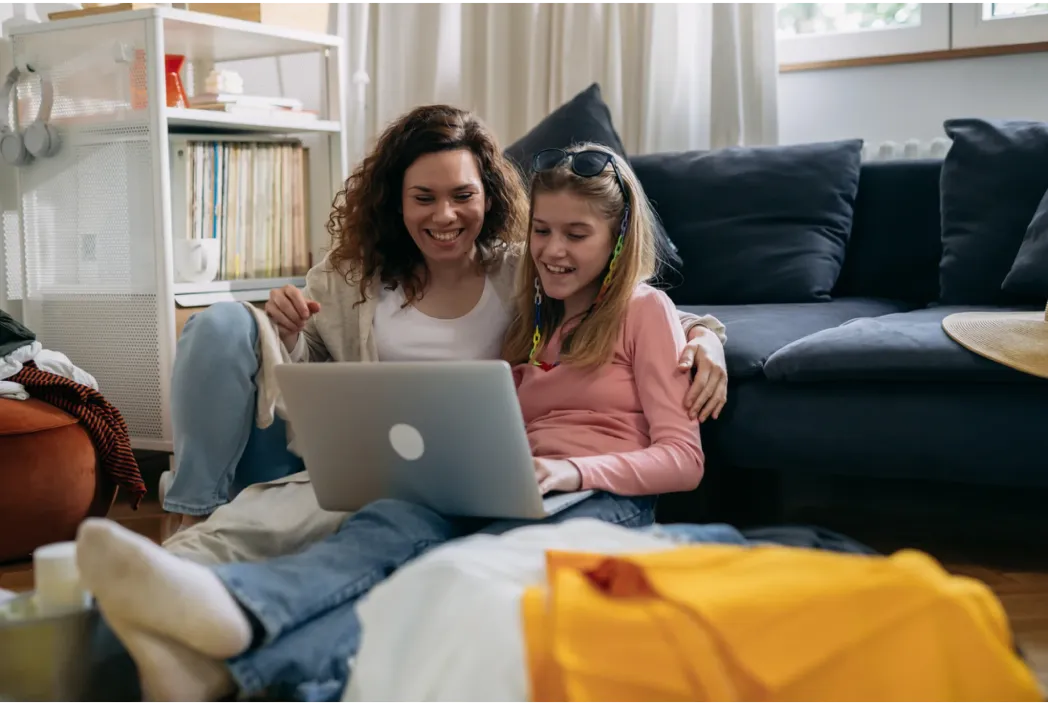 Una mujer y una niña se sientan en el suelo, sonriendo y mirando una computadora portátil. Parecen relajados y felices, rodeados de una decoración interior hogareña e informal.