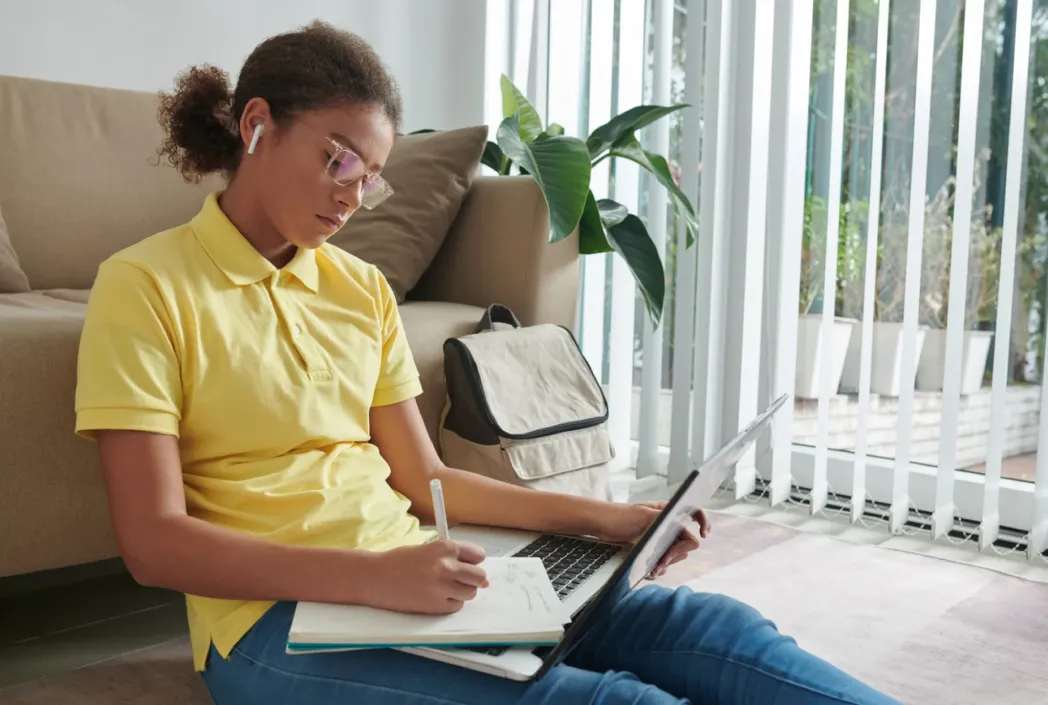 Un joven con cabello rizado y gafas se sienta en el suelo, concentrado en escribir en un cuaderno. Llevan un polo amarillo y auriculares, con una computadora portátil abierta cerca.