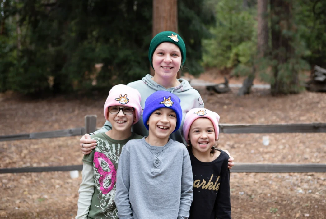Four smiling children wearing colorful beanies stand in front of a wooden fence in a forest setting, conveying a sense of friendship and joy.