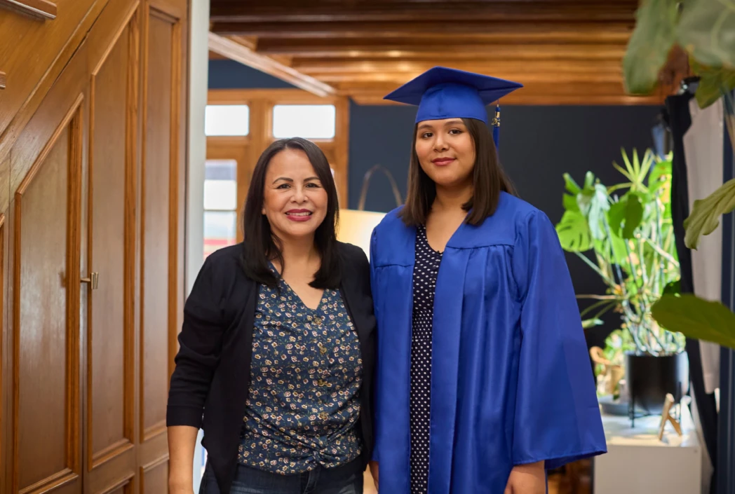 A proud woman stands beside a young graduate in a blue cap and gown, inside a warmly lit home. Plants decorate the background, creating a celebratory mood.