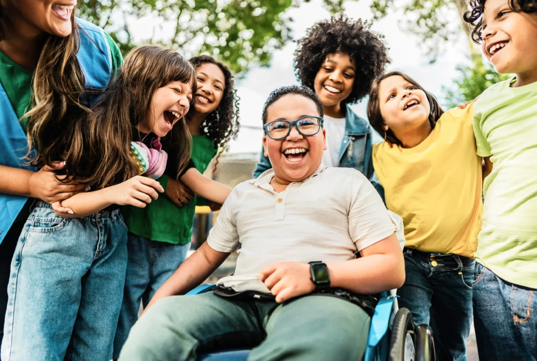 A diverse group of smiling children, one in a wheelchair, share a joyful moment outdoors. The scene conveys friendship, inclusion, and happiness.