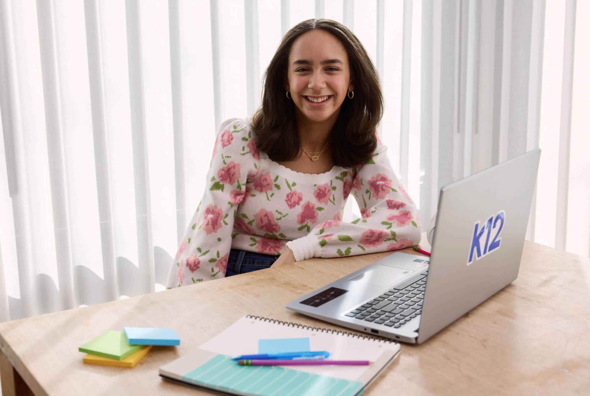 Una adolescente sonriente con un suéter floral se sienta en un escritorio con una computadora portátil etiquetada como "K12", rodeada de un cuaderno, bolígrafos y coloridas notas adhesivas, lo que sugiere aprendizaje en línea o escuela virtual.