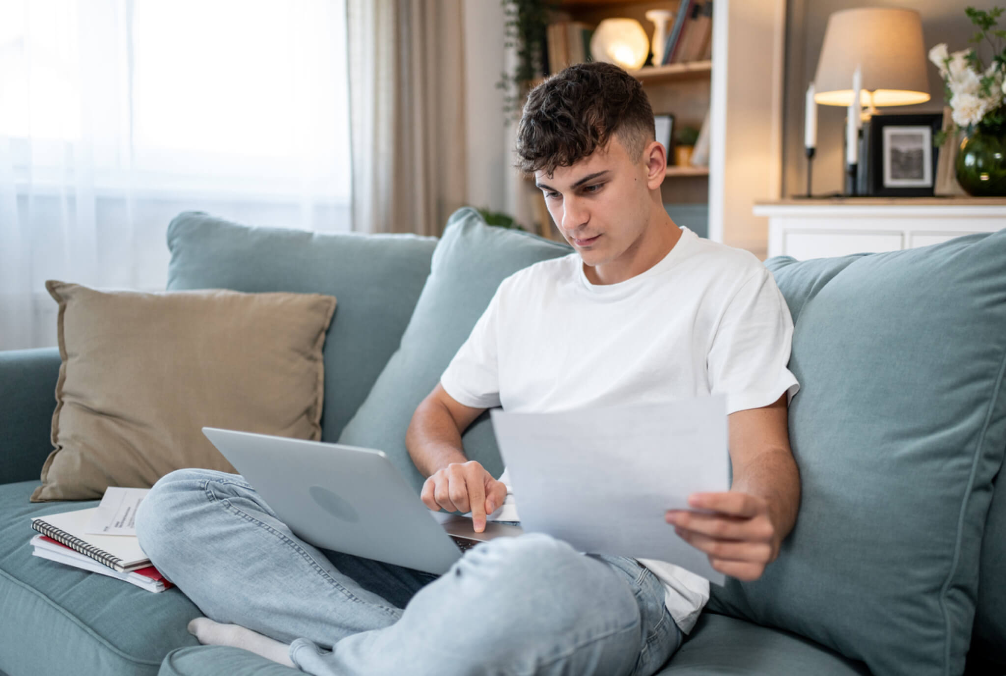 Adolescente sentado en un sofá usando una computadora portátil mientras sostiene una hoja de papel, con cuadernos cerca, enfocado en estudiar o completar la tarea en casa.