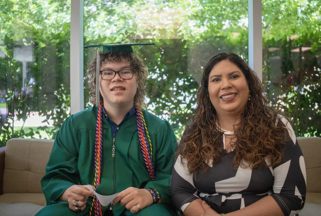 A graduate in a green cap and gown sits smiling beside a woman in a black and white dress. Both seated on a sofa with greenery outside the window.