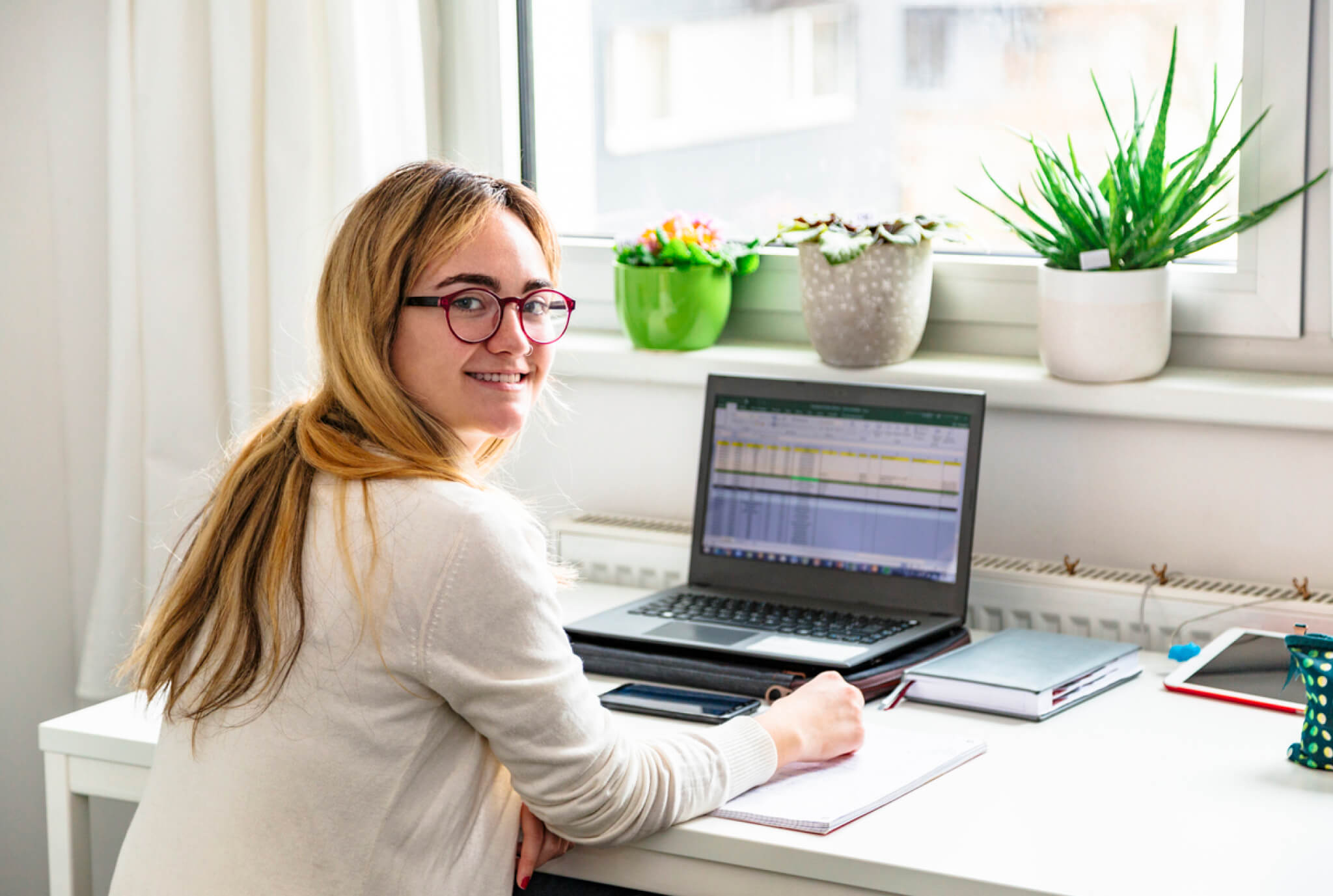 Una mujer joven con cabello largo y rubio y gafas se sienta en un escritorio, sonriendo mientras usa una computadora portátil. Un cuaderno y un teléfono están en el escritorio, con plantas verdes en macetas en el alféizar de la ventana y luz natural que entra por la ventana.