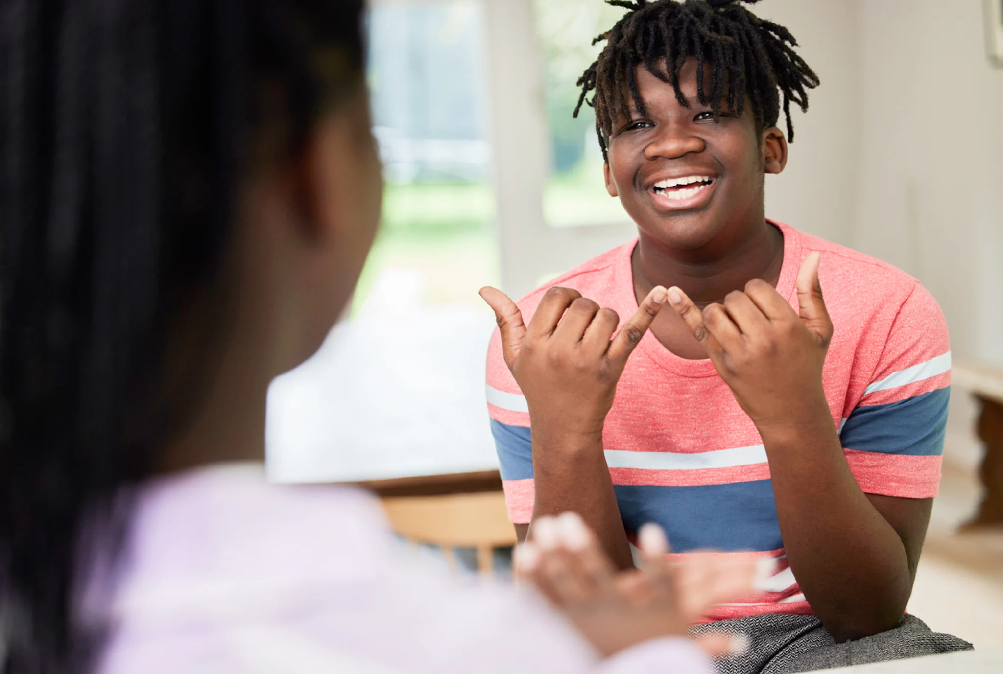 A smiling boy in a striped red shirt communicates using sign language with another person in the foreground. The setting is a bright, indoor space.
