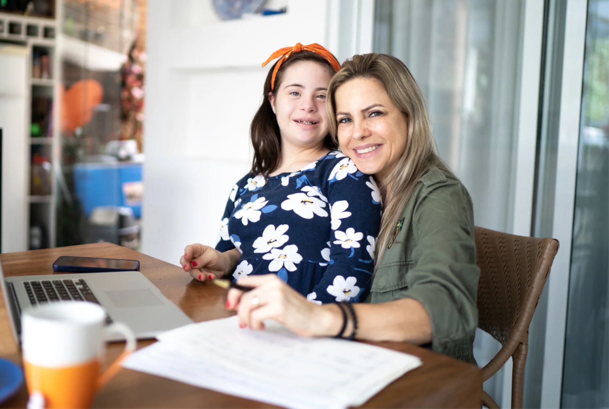 A mother and daughter sit at a table with a laptop and papers, smiling warmly. The daughter wears a floral dress and headband, conveying harmony and support.