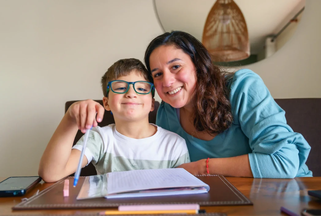 A smiling woman and boy with glasses sit at a table. The boy holds a pen over an open notebook, conveying a positive mood of learning and support.