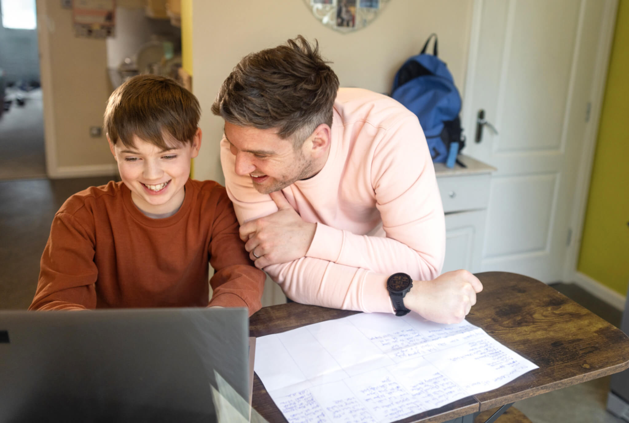 A father and son leaning over a laptop, both smiling and engaged, with a sheet of paper containing handwritten notes on the table, in a cozy kitchen setting.