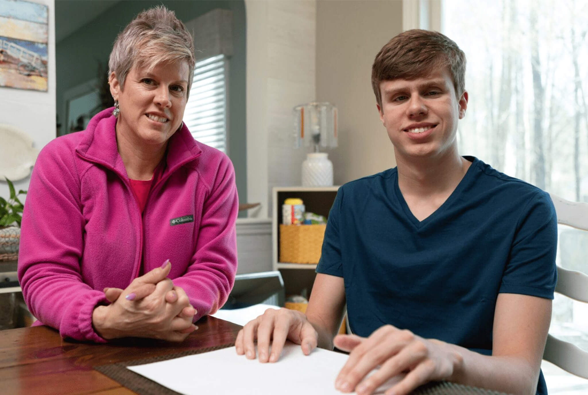 A woman in a pink fleece jacket and a teenage boy in a blue shirt sitting at a table, smiling and looking at the camera, with papers in front of them in a well-lit room.