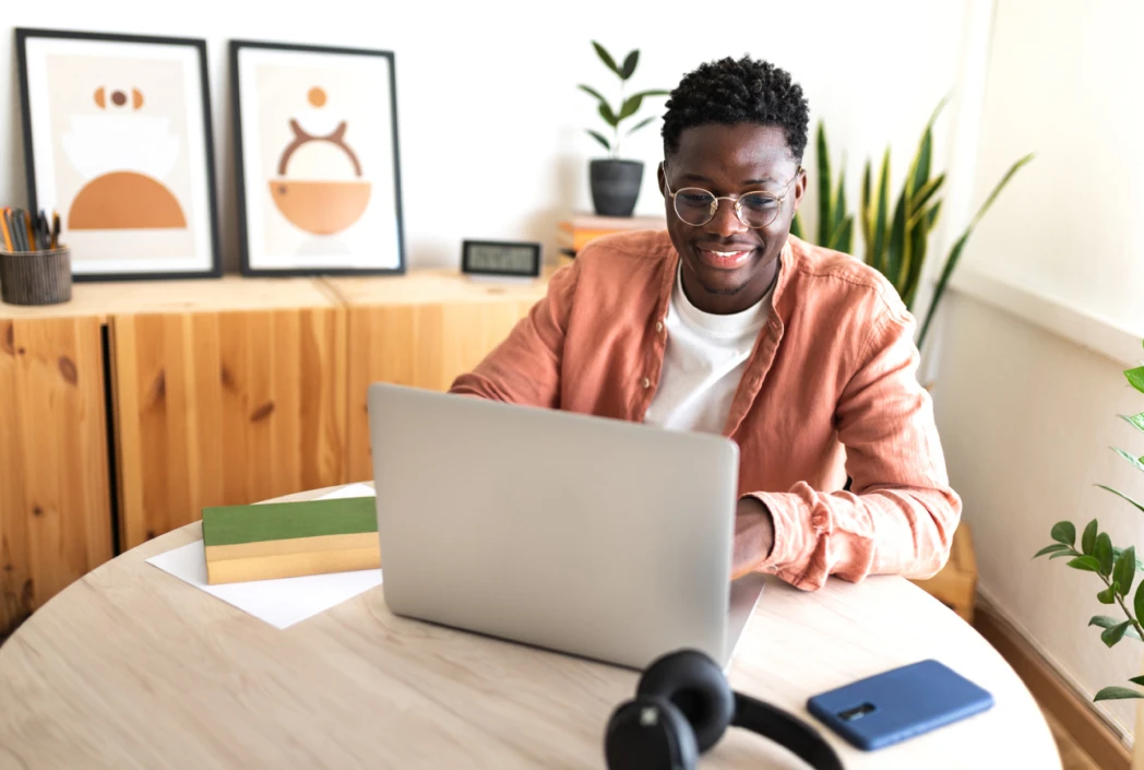 Man in casual orange shirt smiles while working on a laptop at a round table. Nearby are books, headphones, a smartphone, and framed art, with plants adding greenery.