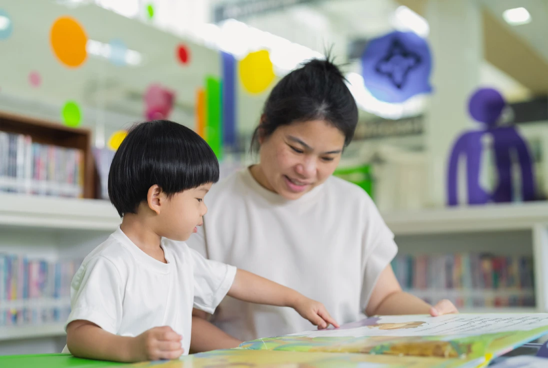 Un niño pequeño y un adulto leyendo un libro ilustrado juntos en una biblioteca. Los colores brillantes y las estanterías crean un ambiente alegre y educativo.
