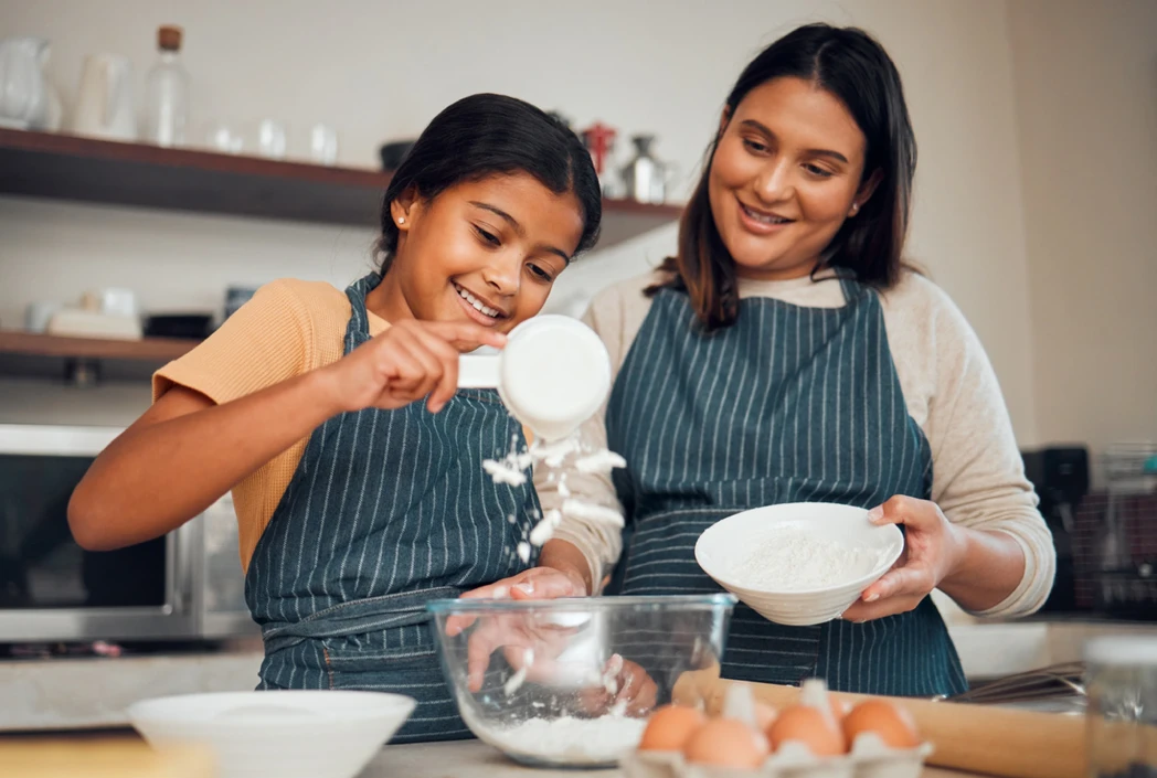 Una mujer sonriente y una niña, ambas con delantales a rayas, horneando alegremente. La niña vierte harina en un tazón para mezclar en un mostrador de la cocina con huevos.