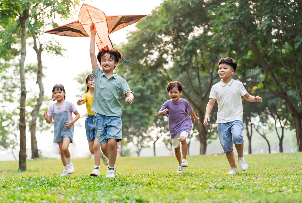 Cinco niños alegres corren por un parque cubierto de hierba, con un niño a la cabeza, sosteniendo una cometa sobre su cabeza. La escena es animada y despreocupada.
