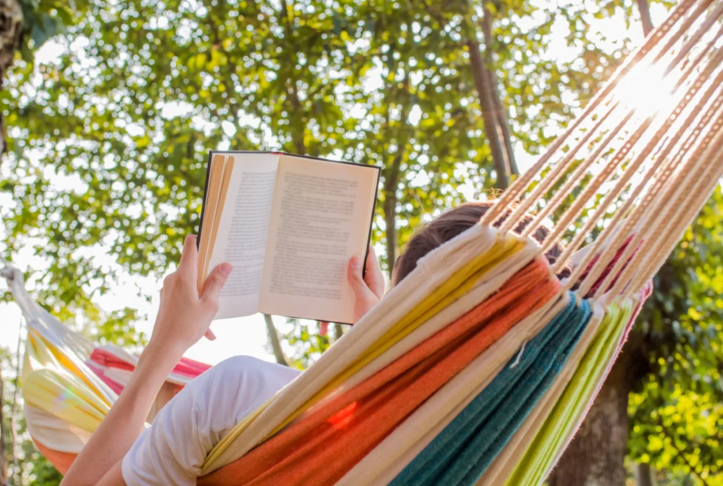 Person relaxing in a colorful hammock under trees, reading a book. Sunlight filters through leaves, creating a serene, peaceful atmosphere.