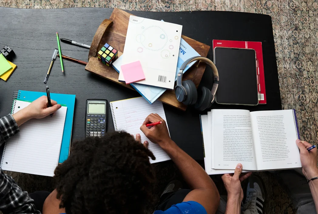 Two people studying at a table with notebooks, a textbook, calculator, headphones, pens, sticky notes, and a Rubik's Cube. The setting feels focused.